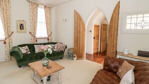Een woonkamer met een groene bank, een bruine leren bank en een houten salontafel. Er zijn bloemen gordijnen en een fotolijst aan de muur.
