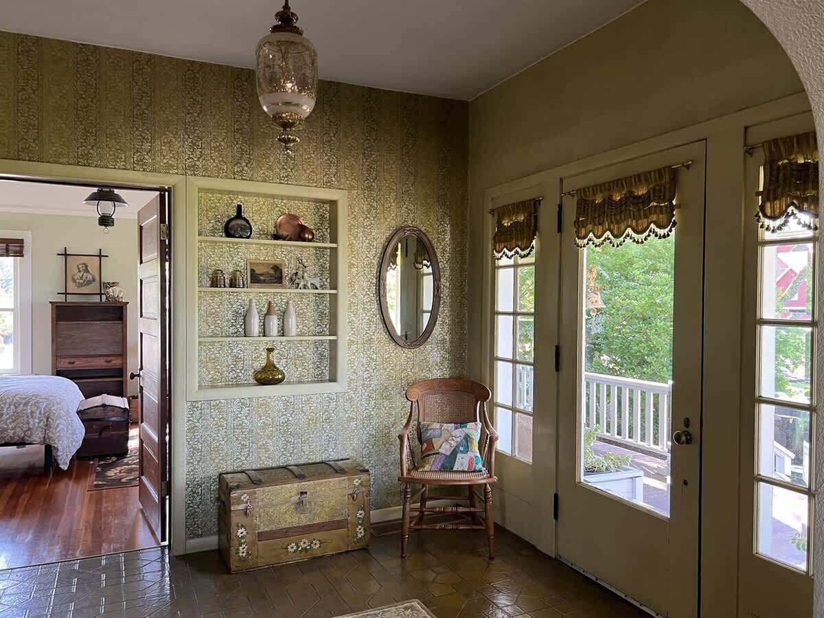 Een kamer met een houten vloer, een bed, een stoel, een dressoir, een spiegel en een raam met uitzicht op groen.