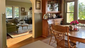 A cozy living room with a sofa, coffee table, and a dining area with a table and chairs. There is a kitchen area with cabinets and a window. - Barn Owl Hill - Historic Farmhouse Vineyard Views (Rickreall)