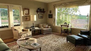 A spacious living room with a large window, a patterned rug, and a variety of seating options including a sofa, armchairs, and a coffee table. - Barn Owl Hill - Historic Farmhouse Vineyard Views (Rickreall)