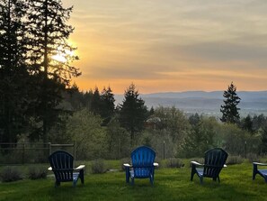 Three Adirondack chairs facing a sunset over a grassy field with trees and distant mountains.