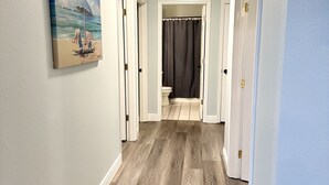 A hallway with wooden flooring, a painting of a beach scene, and a bathroom visible through an open door. - "Bayou Breeze" Private Pool (Heated) Gorgeous Home! Bikes, Game room (Navarre)