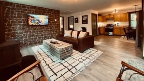 A living room with a stone wall, a fireplace, a sofa, and a coffee table. There is a kitchen in the background with wooden cabinets and a dining area.