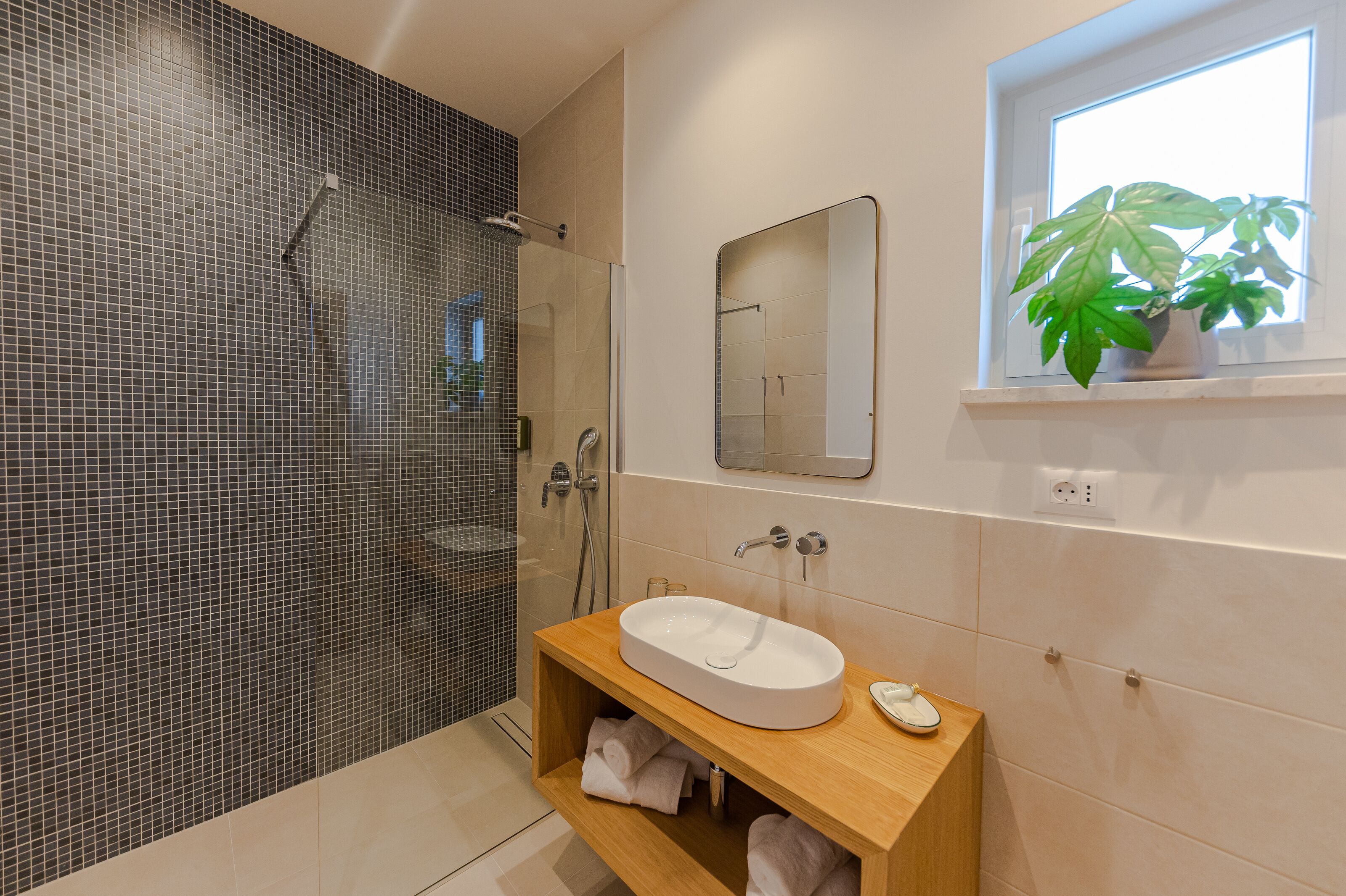 A modern bathroom with a glass-enclosed shower, a wooden vanity with a sink, and a potted plant by the window.