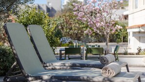 A poolside area with lounge chairs and a table, surrounded by greenery and a building in the background. - The Olive Poolhouse (Split)