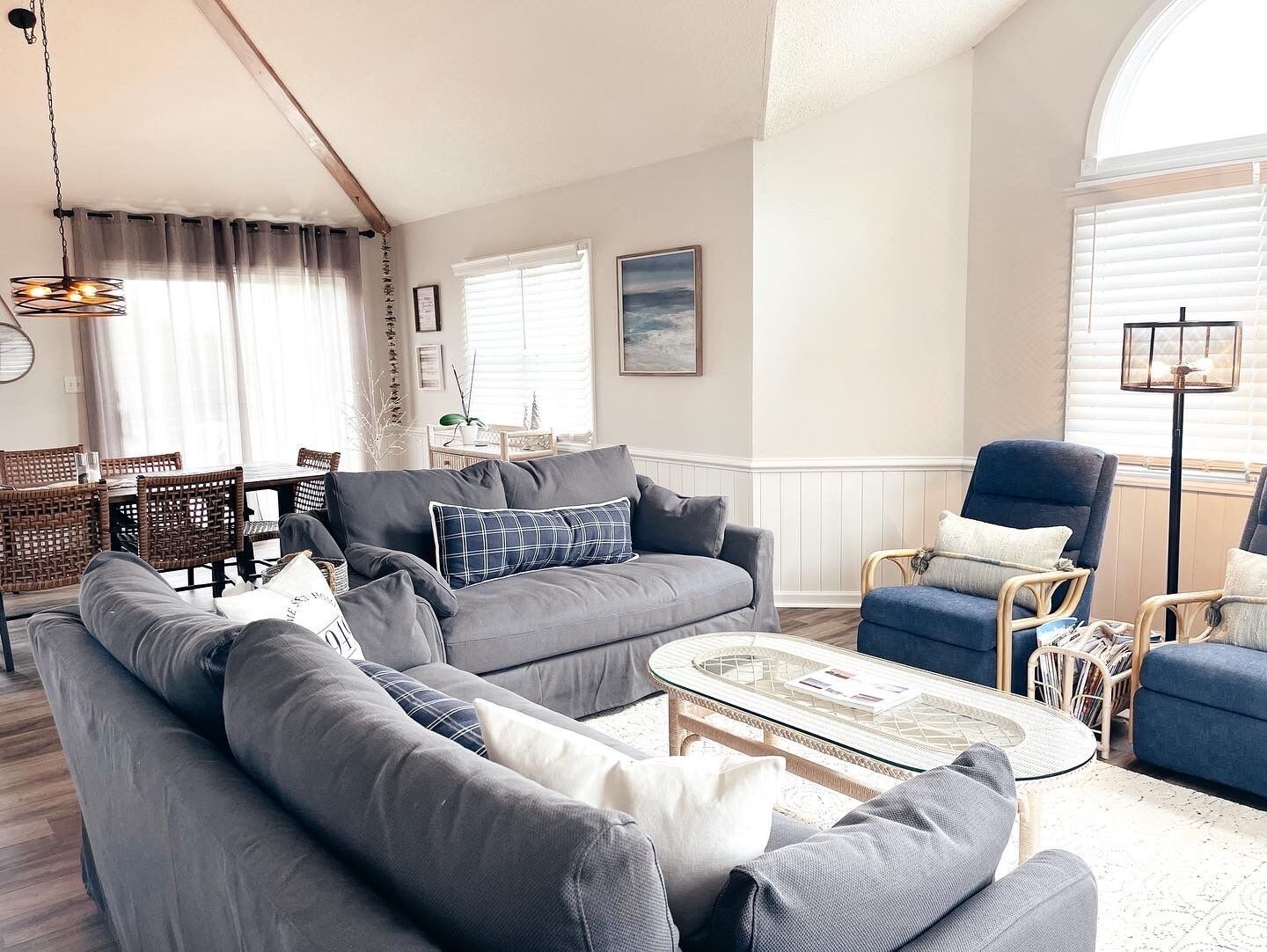 Een woonkamer met een grijze bank, een blauwe fauteuil, rieten stoelen en een glazen salontafel.