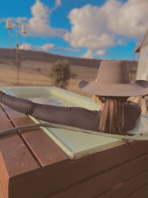 Una persona sentada en un banco de madera al aire libre, con sombrero, pantalones largos y una blusa de manga larga.