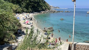 A beach with clear blue water, people swimming and sunbathing, and a rocky hillside in the background.