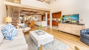 A spacious living area with a sofa, coffee table, and a television on a wooden cabinet. A staircase leads to an upper level, and the room has wooden flooring and a ceiling fan. - Breathtaking Ocean Views Kahana Village 40 2BR/2BA Condo Pool, Beach Access (lahaina)