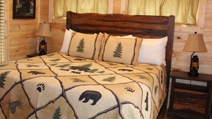 Ein Schlafzimmer einer Holzhütte mit einem Bett, das eine Bären-Quiltdecke hat, zwei Nachttischlampen und einem Fenster mit Vorhängen.