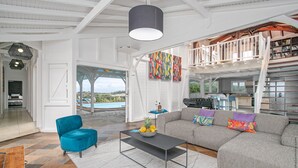 Een ruime woonkamer met een grijze bank, een blauwe fauteuil en een salontafel. Door een open deur is een balkon met uitzicht op een zwembad te zien.