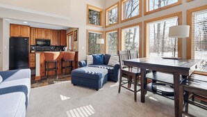 A spacious kitchen with wooden cabinets, a black refrigerator, and a microwave. There is a dining area with a table and chairs, and a blue sofa with a patterned rug. Large windows with blinds provide natural light. - Ski In/Out| Combined Upper and Lower Unit 780/781 | 5 Bedroom | Gas Fireplace (Boyne Fallss)