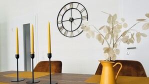 A dining area with a wooden table, yellow placemats, a yellow vase with white flowers, and a black wall clock.