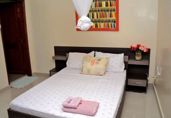 A neatly made bed with a floral-patterned bedspread and pink towels, a bedside table with a vase of flowers, and a colorful abstract painting on the wall.
