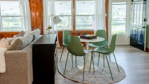 Een gezellige woonkamer met een bank, een salontafel en een schaakbord. Er zijn grote ramen met gordijnen, een plafondventilator en een deur met een ruit.