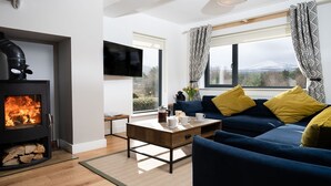A modern living room with a wood-burning stove, a sofa with yellow cushions, and a large window with a view. - Two Queens Houses -  a house that sleeps 10 guests  in 5 bedrooms (Aviemore)