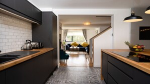 A modern kitchen with dark cabinetry, a wooden countertop, and a patterned backsplash. There is a dining area with a sofa and a coffee table, and a fruit bowl on the counter. - Two Queens Houses -  a house that sleeps 10 guests  in 5 bedrooms (Aviemore)