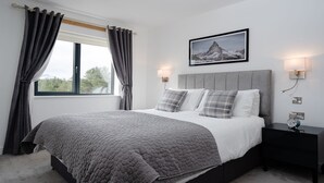 A bedroom with a large bed, grey curtains, a framed picture on the wall, and bedside lamps. - Two Queens Houses -  a house that sleeps 10 guests  in 5 bedrooms (Aviemore)