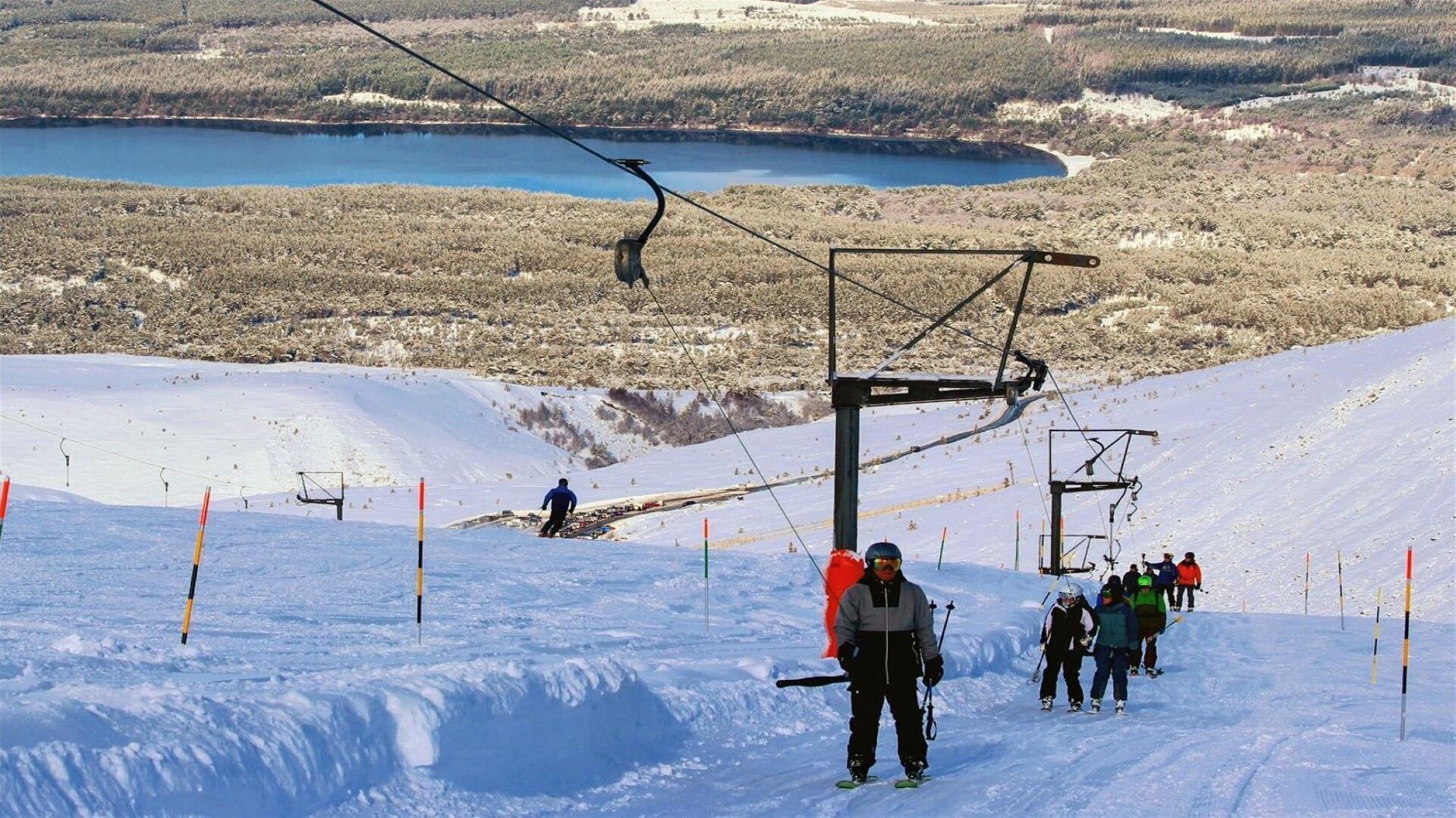 Een skilift met skiërs op een besneeuwde helling, een helder uitzicht op een meer en een bebost gebied op de achtergrond.