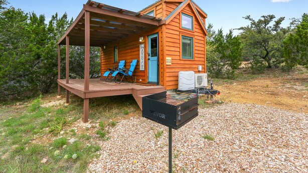 A wooden cabin with a covered porch, two blue lounge chairs, and a grill. - Pet-Friendly Tiny Home in Dripping Springs w/ Hill Country Views (Dripping Springs)