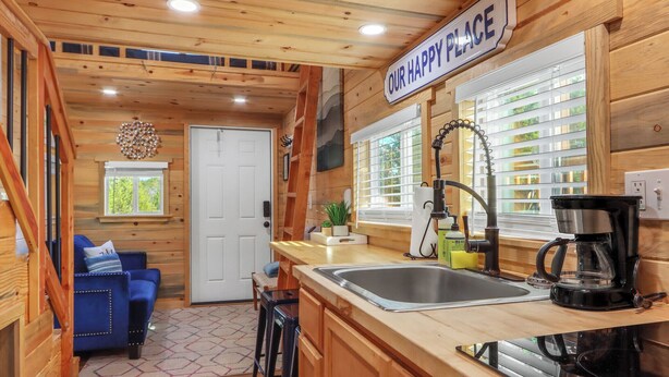 A cozy cabin interior with a wooden interior, a kitchen area with a sink and coffee maker, a blue sofa, and a sign reading "OUR HAPPY PLACE". - Cozy Tiny House in Dripping Springs w/ Sunset Views & Romantic Fireplace (Dripping Springs)