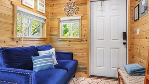 A cozy room with a blue sofa, two windows with blinds, a white door, and a wooden ceiling. - Cozy Tiny House in Dripping Springs w/ Sunset Views & Romantic Fireplace (Dripping Springs)
