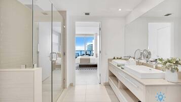 A modern bathroom with a glass shower enclosure, a large mirror, and a view of a bedroom with a bed and a cityscape.