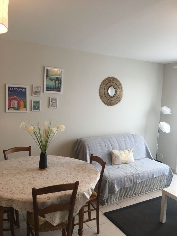 A dining area with a round table, chairs, a vase with flowers, and a sofa with a blanket. There are framed pictures on the wall and a decorative mirror.
