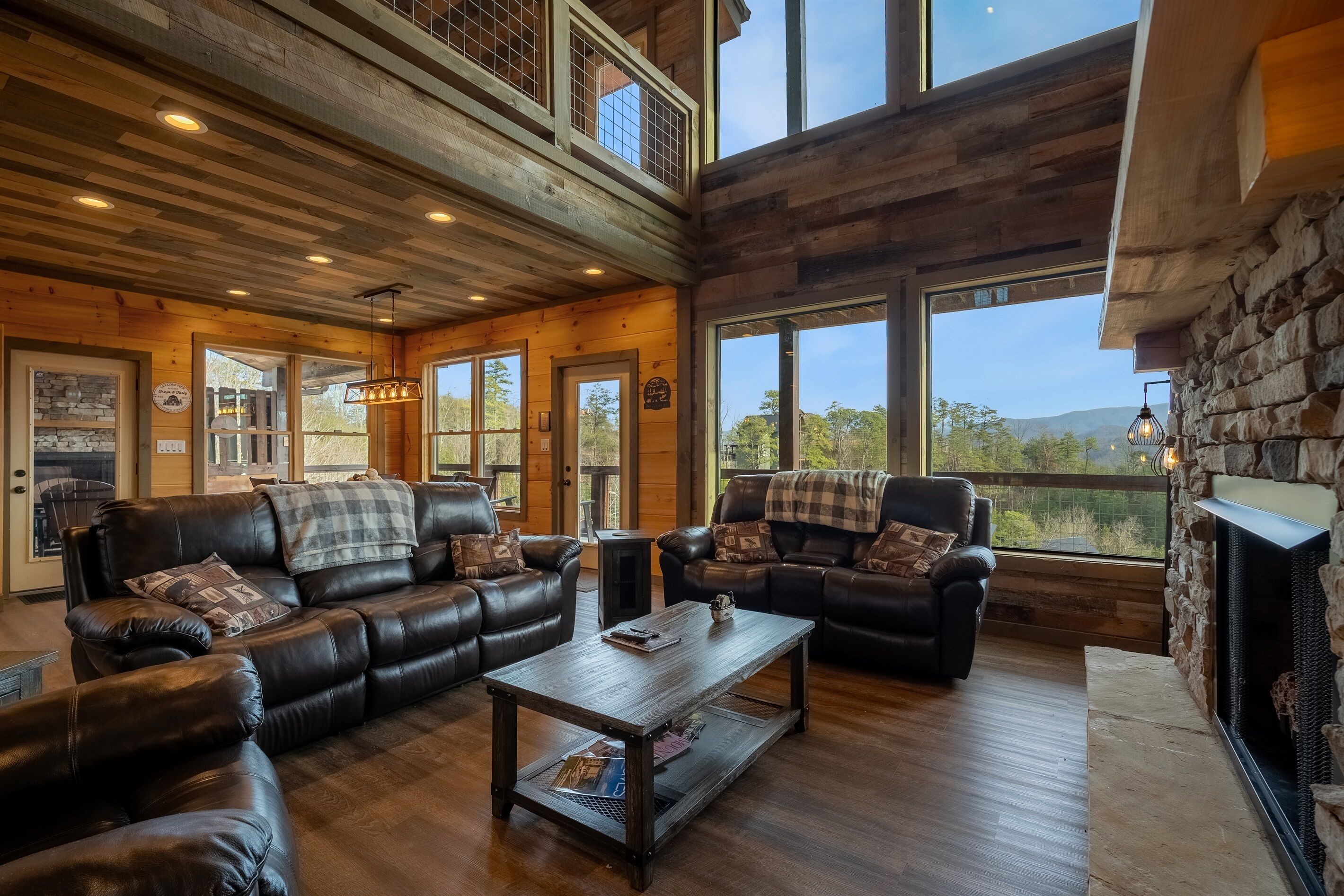A spacious living room with leather sofas, a coffee table, and a stone fireplace. Large windows offer a view of the outdoors.