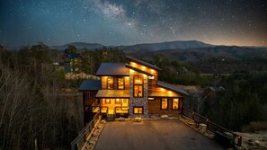Una cabaña grande de troncos de varios pisos, con base de piedra, rodeada por un muro de piedra y una terraza de madera.