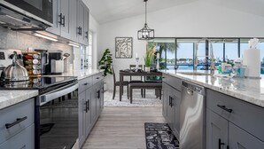 A modern kitchen with grey cabinets, stainless steel appliances, and a view of the marina through large windows. - 295 Ostego Dr - Gorgeous Bay Front Home 4 Bedroom Home by RedAwning (Fort Myers Beach)