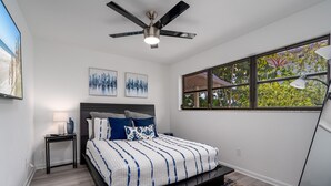 A bedroom with a bed, a ceiling fan, a nightstand, a lamp, a window with a view of trees, and artwork on the wall. - 295 Ostego Dr - Gorgeous Bay Front Home 4 Bedroom Home by RedAwning (Fort Myers Beach)