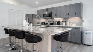 A modern kitchen with grey cabinets, a stainless steel microwave, a large island with bar stools, and a trash can. - 295 Ostego Dr - Gorgeous Bay Front Home 4 Bedroom Home by RedAwning (Fort Myers Beach)