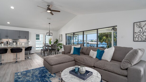 A modern living room with a sectional sofa, a round coffee table, and a view of the ocean through large windows. - 295 Ostego Dr - Gorgeous Bay Front Home 4 Bedroom Home by RedAwning (Fort Myers Beach)