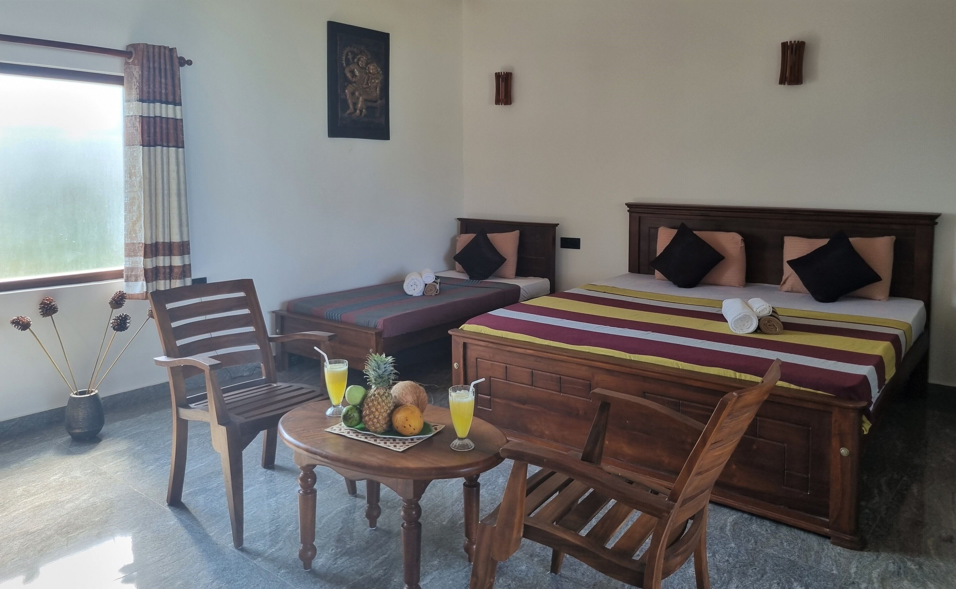 A hotel room with two single beds, a wooden table with fruits and a drink, a chair, and a window with curtains.