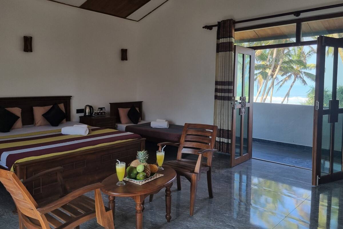 A hotel room with two beds, a wooden table with fruit and drinks, a chair, and a view of palm trees through a glass door.
