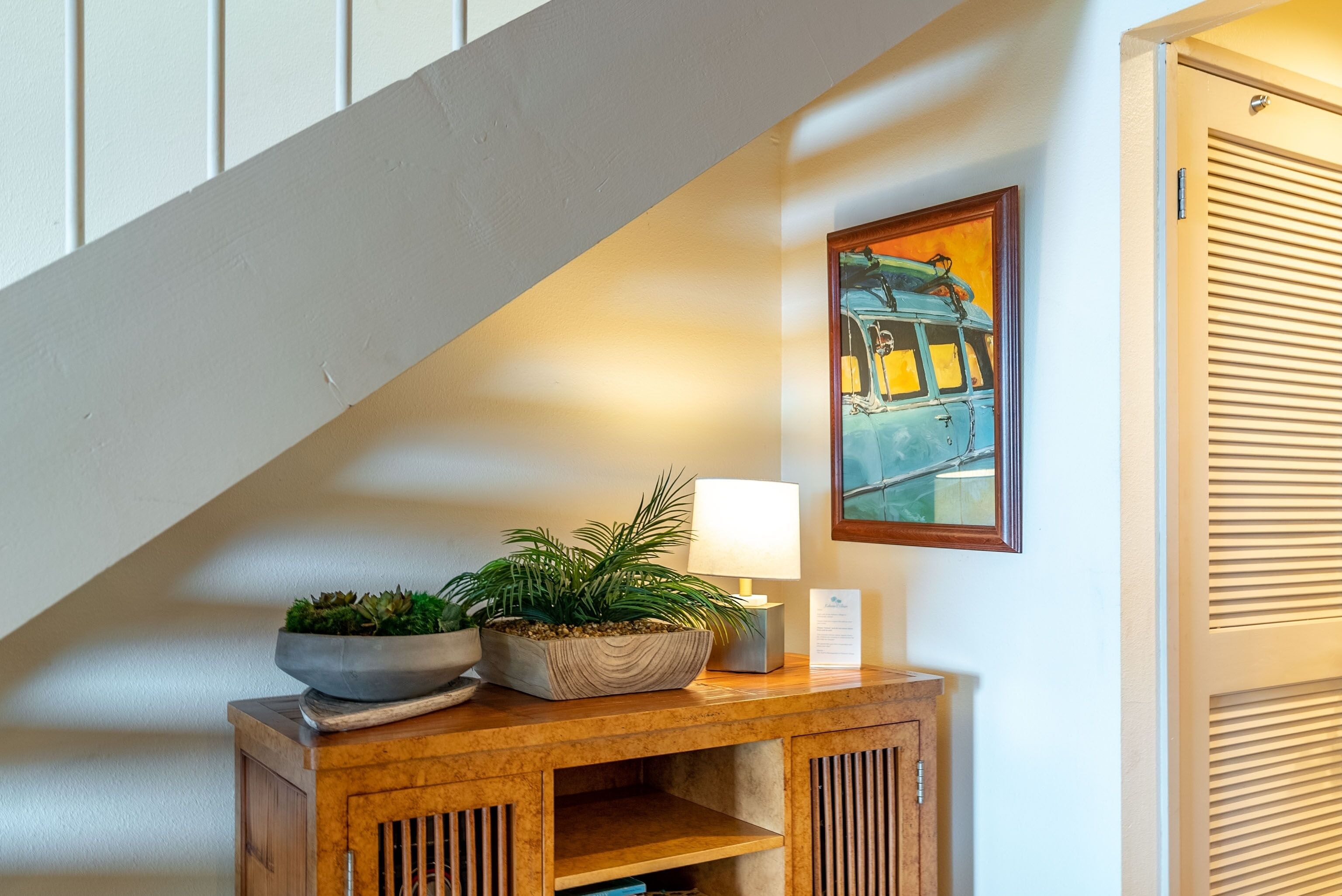 A wooden console table with a potted plant and a lamp, a painting of a vintage car on the wall, and a staircase in the background.
