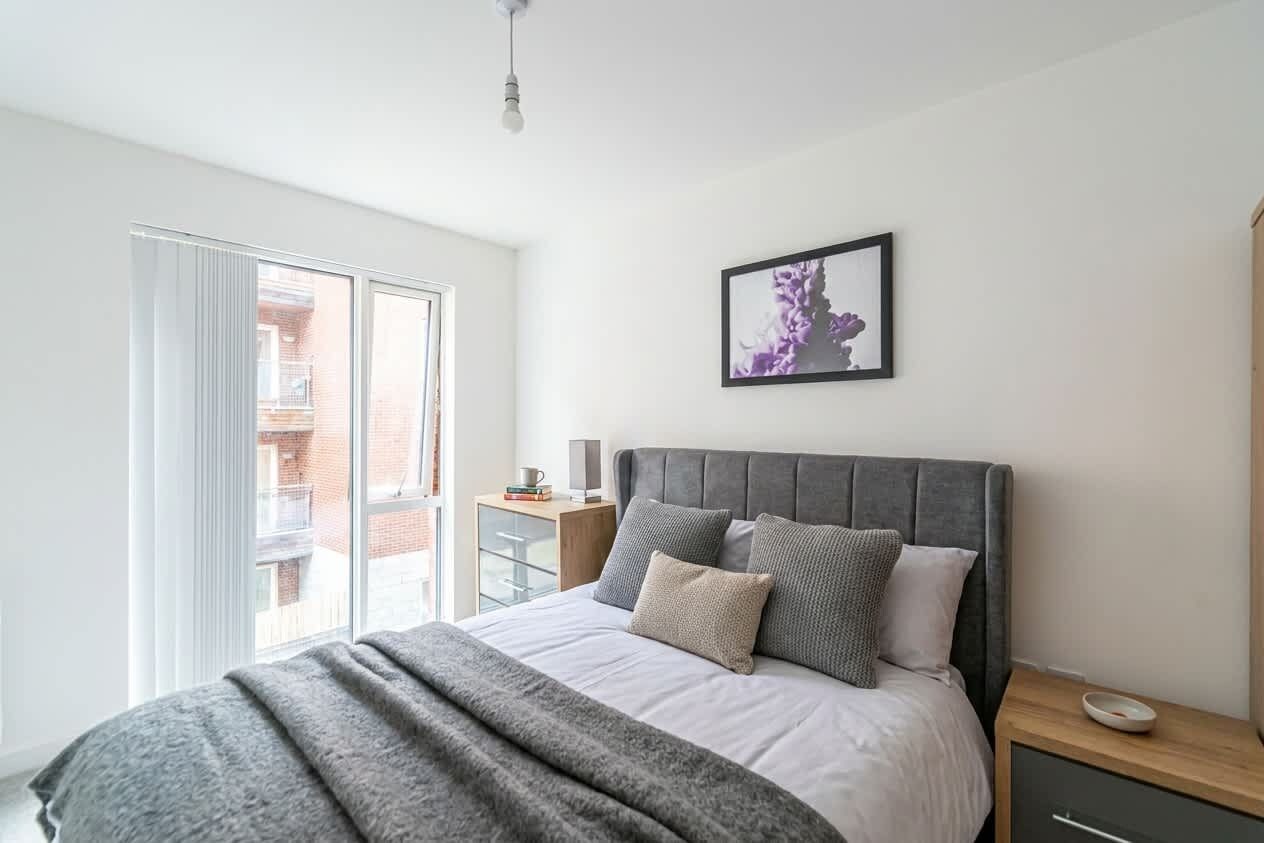 A bedroom with a bed, a bedside table, a nightstand, a framed picture on the wall, and a window with a view of a building outside.