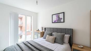 A bedroom with a bed, a bedside table, a nightstand, a framed picture on the wall, and a window with a view of a building outside.