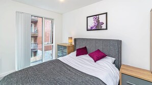 A bedroom with a bed, a nightstand, a lamp, a framed picture, and a sliding door to a balcony. - Central Manchester 2BR Apartment + Free Parking (Manchester)