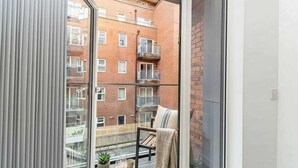 Un balcon donnant sur un immeuble en briques, une chaise, une petite table avec une plante et une fenêtre avec des stores.