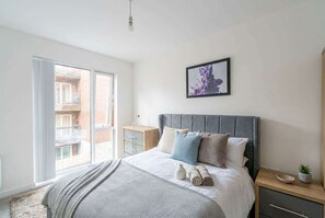Un dormitorio con cama, mesita de noche, ventana y un cuadro enmarcado en la pared.