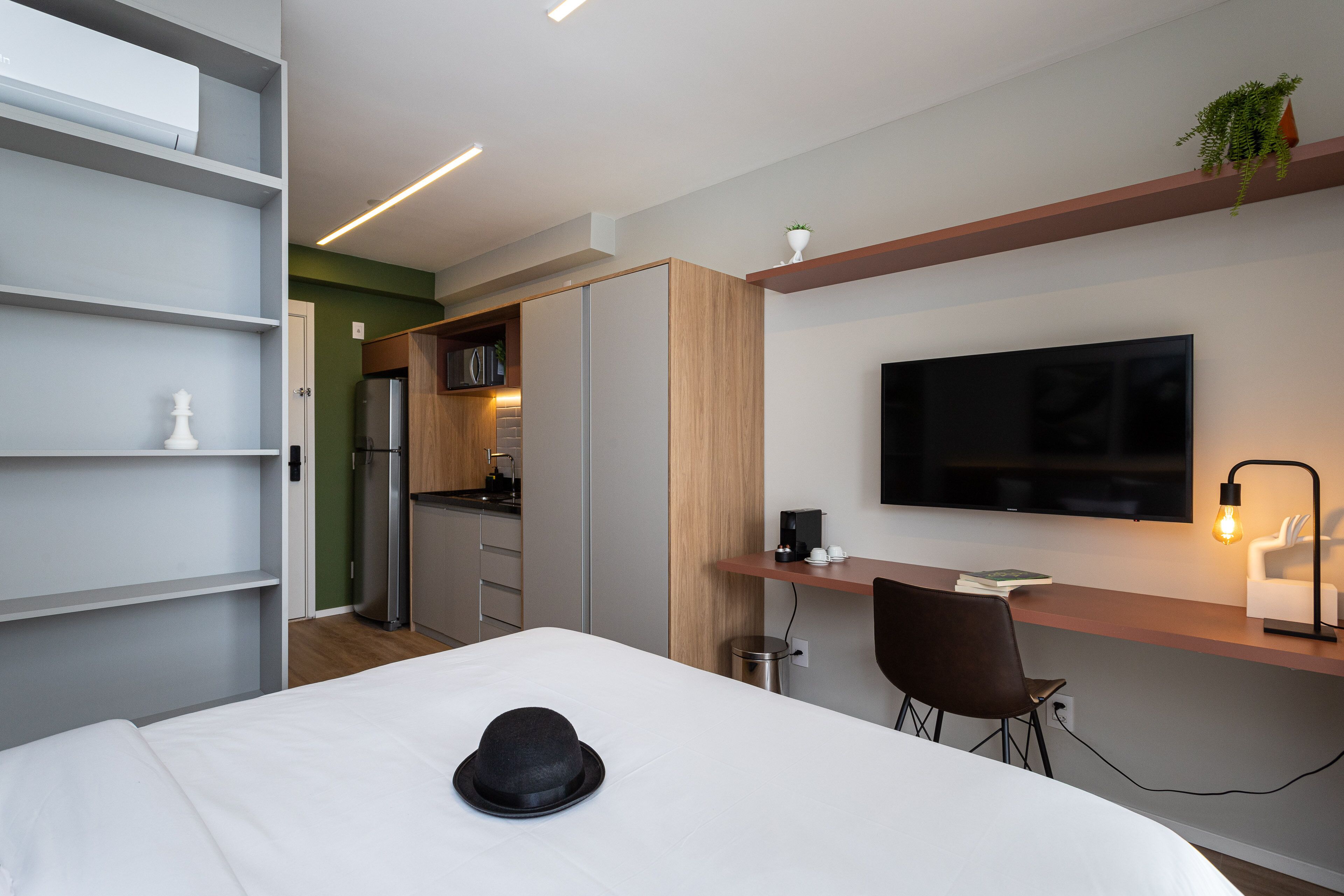 A modern hotel room with a bed, a desk, a chair, a TV, and a kitchenette.