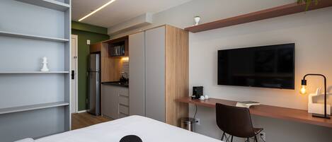 A modern hotel room with a bed, a desk, a chair, a TV, and a kitchenette.