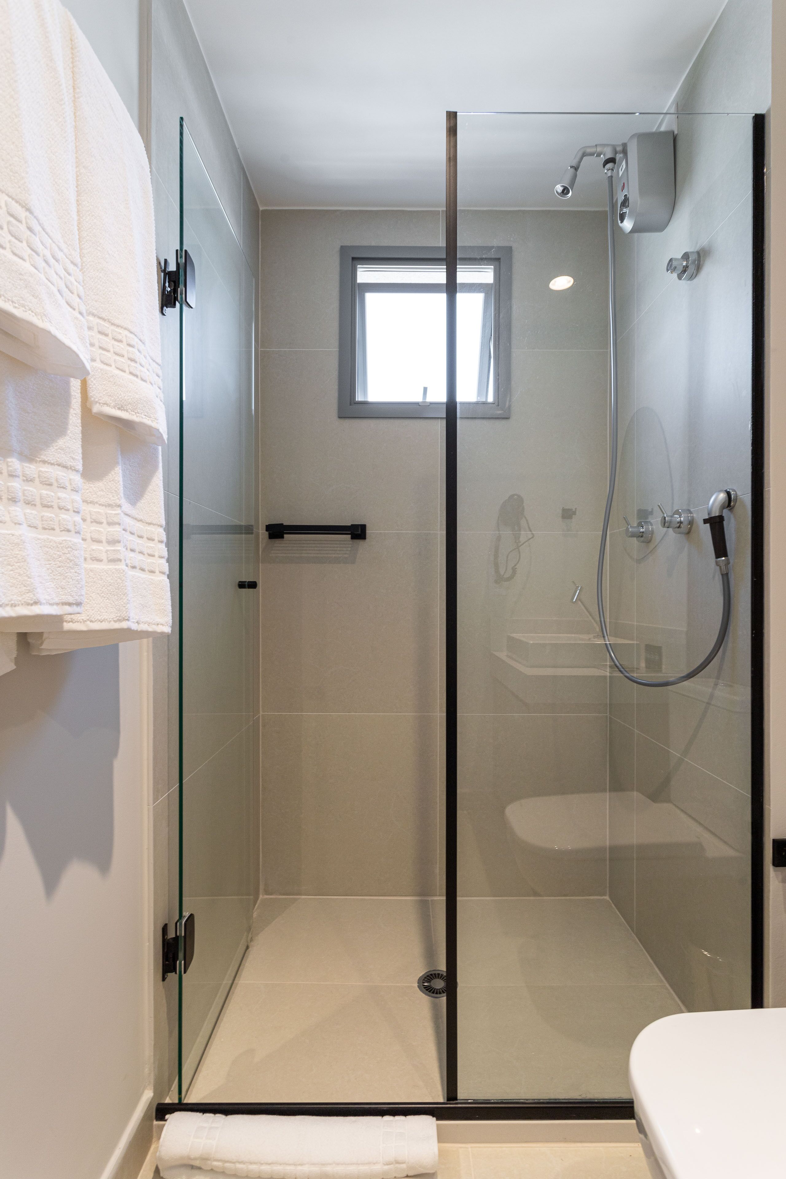 Une douche fermée par des parois de verre, dotée d’un pommeau de douche à main, d’une porte en verre et d’une petite fenêtre située au-dessus de la douche.