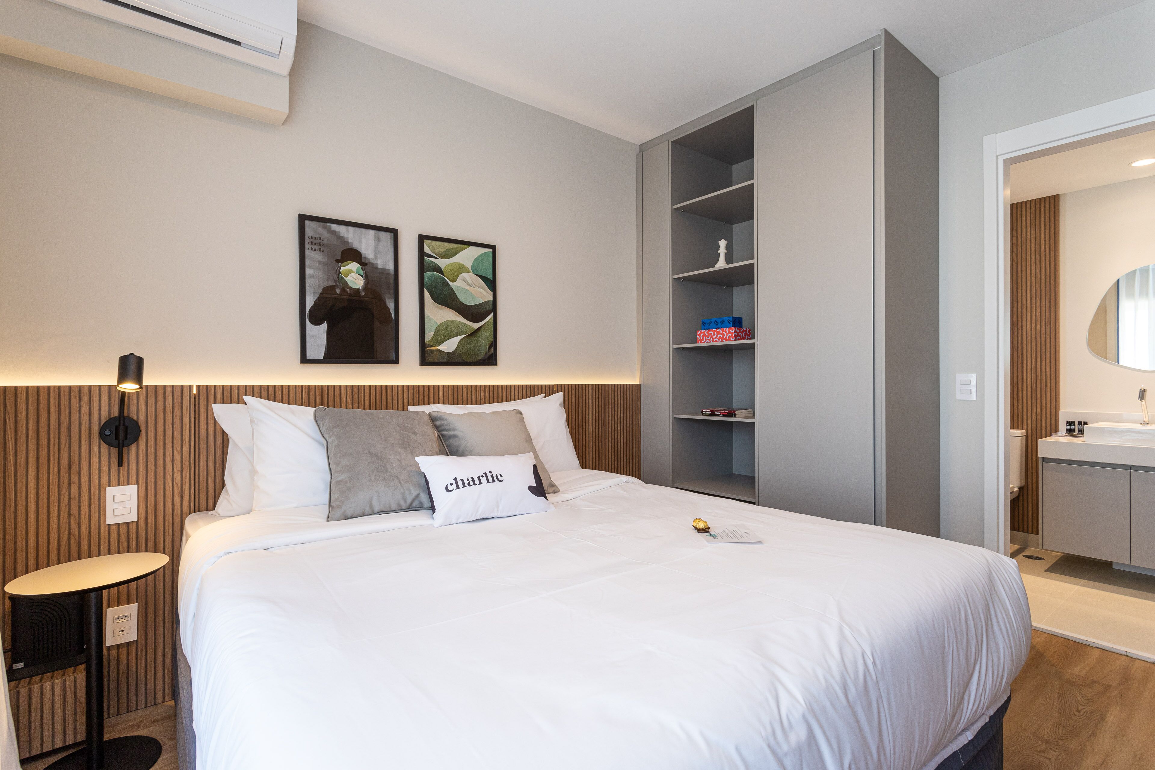 A modern hotel room with a large bed, a bedside table, a built-in wardrobe, and a bathroom with a mirror and sink.