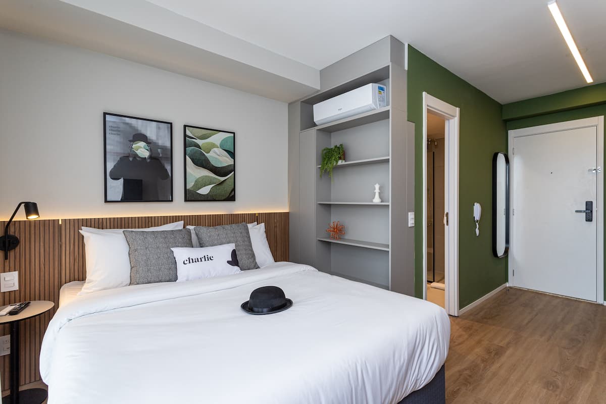 Una habitación de hotel moderna con una cama grande, una mesita de noche con lámpara, aire acondicionado de pared y una puerta que da al baño.