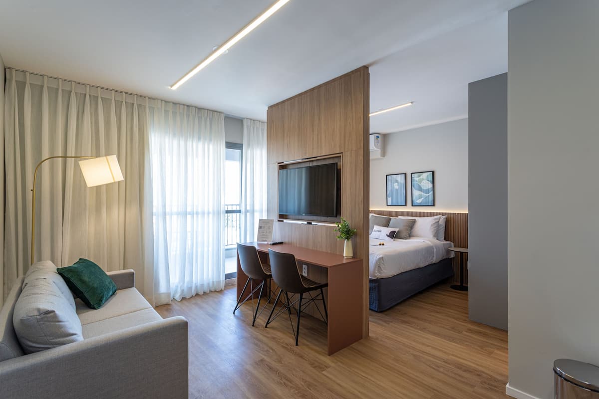 Una habitación de hotel moderna con sofá, escritorio con dos sillas, cama, televisor y un amplio ventanal con cortinas.