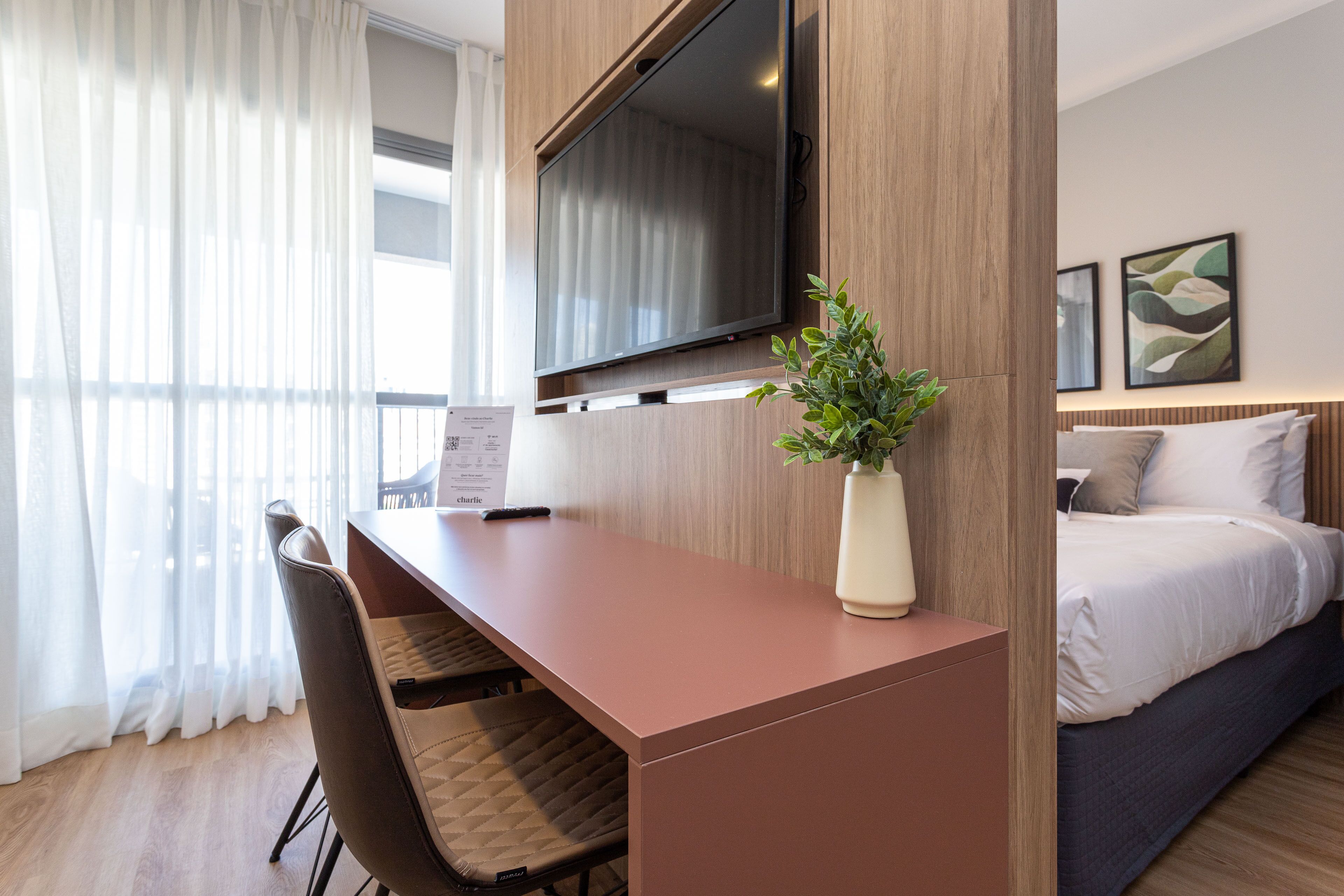 A modern hotel room with a wooden desk, a flat-screen TV, a bed with white bedding, and a potted plant.