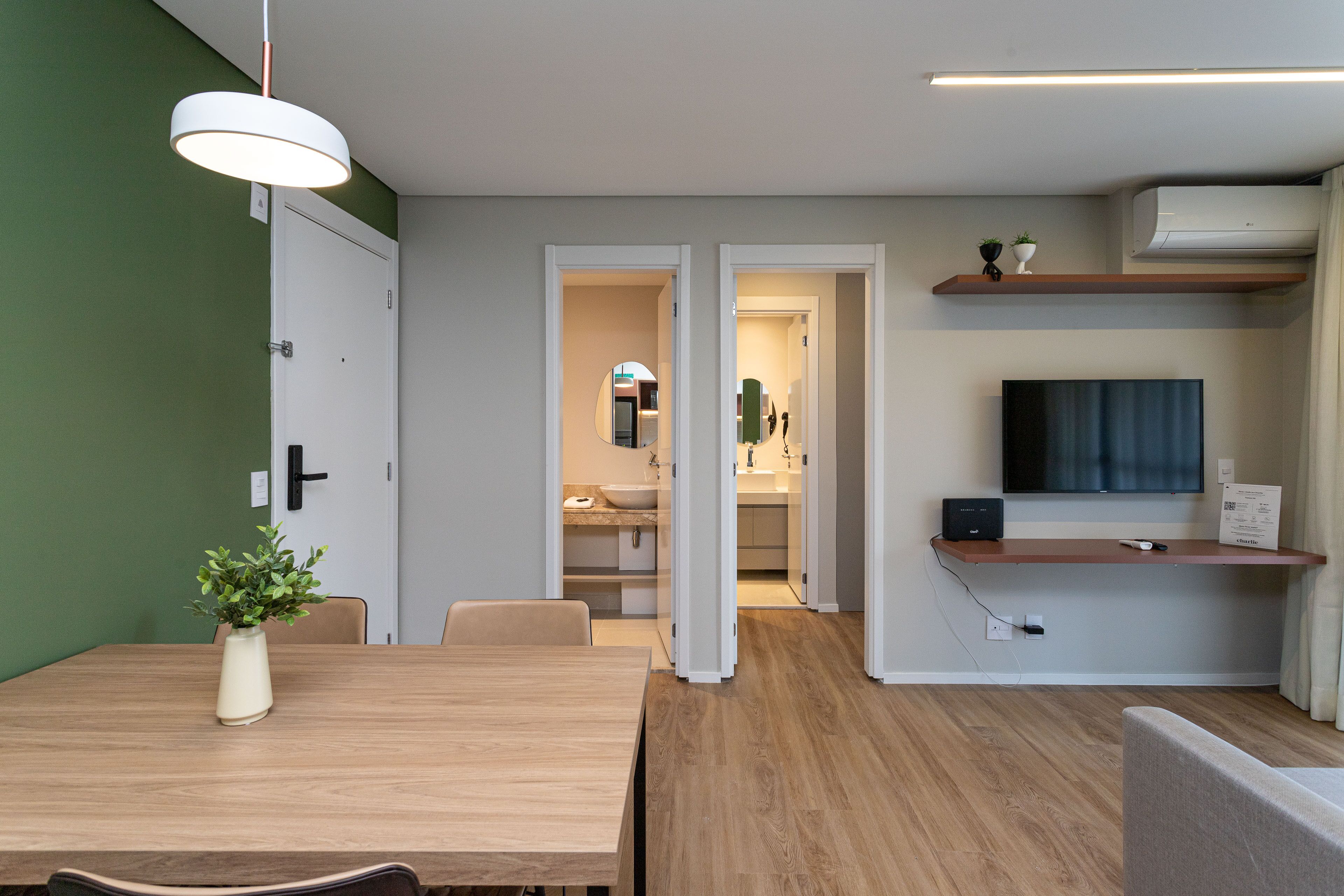 A modern living room with a wooden dining table, a potted plant, a TV on a shelf, and a kitchen in the background.
